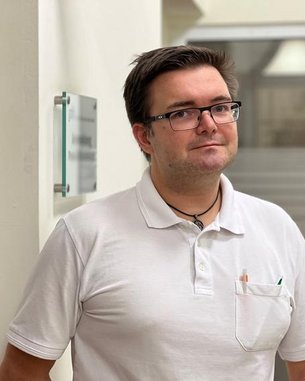Ein Mann mit Brille und einem weißen Polohemd steht an einer Wand. Er hat einen Stift in der Brusttasche und schaut in die Kamera.