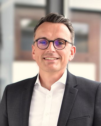 Ein Mann mit Brille und einem Anzug steht lächelnd vor einem hellen Hintergrund. Er hat kurzes, graues Haar und trägt ein weißes Hemd unter dem Anzug.