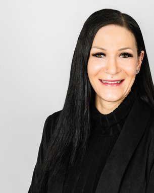 Eine Frau mit langen, schwarzen Haaren und einem schwarzen Blazer lächelt in die Kamera vor einem neutralen Hintergrund.