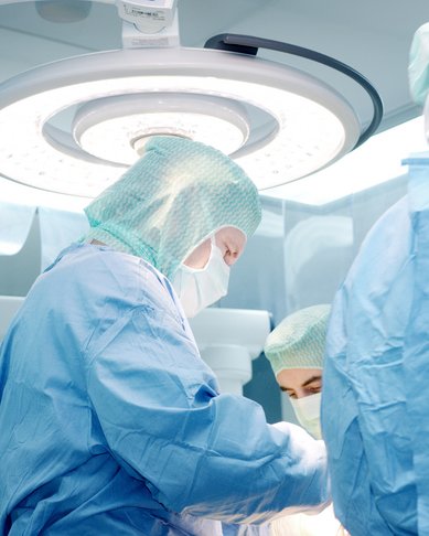 Chirurgen in blauer OP-Kleidung führen einen Eingriff durch. Sie tragen Schutzmasken und Hauben. Ein helles Operationslicht beleuchtet den Raum, während sie konzentriert an einem Patienten arbeiten.