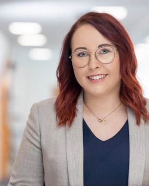 Eine Person mit roten Haaren und Brille trägt einen grauen Blazer über einem dunklen Oberteil. Sie lächelt freundlich und steht in einem modernen, hellen Raum mit unscharfem Hintergrund.