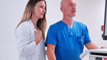 Zwei medizinische Fachkräfte, eine Frau in einem weißen Kittel und ein Mann in einem blauen Scrub, stehen an einem medizinischen Gerät und sprechen miteinander.