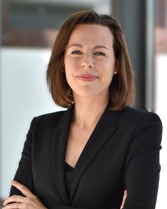 Eine Frau mit schulterlangen braunen Haaren trägt einen schwarzen Blazer und steht mit verschränkten Armen. Sie blickt direkt in die Kamera und hat einen neutralen Gesichtsausdruck.